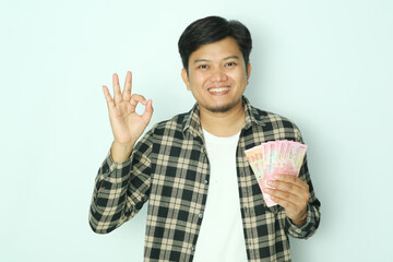 Young Asian man wearing brown flannel shirt smiling while holding wallet with money and give OK finger sign