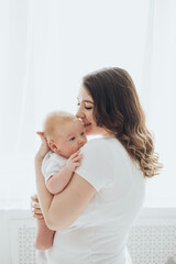 A happy young mother and a newborn baby smile, hug and kiss on a white isolated window background, a loving family at home