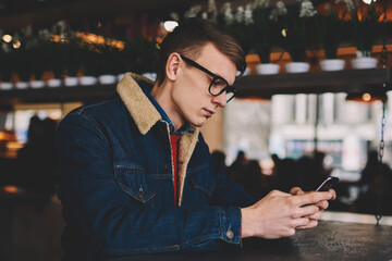 Pensive hipster blogger chatting online with followers on smartphone connected to 4G internet sitting in coffee shop.Young man in denim jacket installing application on telephone resting in cafe - Powered by Adobe