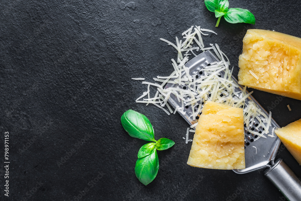 Poster Parmesan cheese slices with basil leaves and grater on a dark stone background with copy space top view