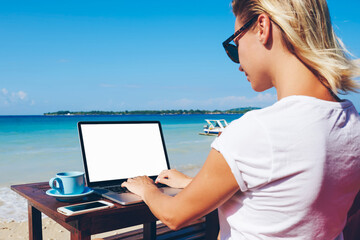 Young woman millennial keyboarding on netbook with mock up screen communicating in online chat sitting near ocean, businesswoman working distantly on vacation in tropical environment enjoying nature