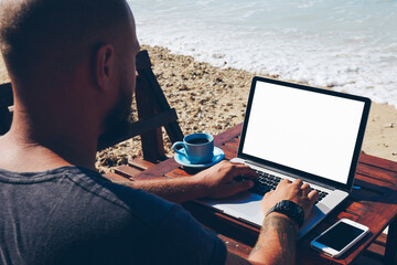 Back view of skilled male freelancer doing distance job on ocean shore browsing information on...