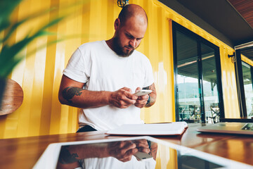 hipster guy chatting in social networks via mobile phone sending text message, young millennial male holding smartphone for installing new application standing near desktop in office with devices