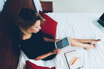 Top view of female blogger reading infrormation from social media holding portable pc lying on bed,...