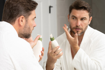 A man is using a mirror to inspect his nose, eyes, jaw, and beard while holding a bottle of lotion...