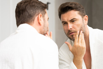 a man in a white bathrobe in the bathroom looking at the skin on his faceSpa male model, skincare...