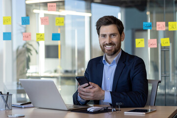 Portrait of successful man in business suit in office interior, mature businessman smiling and looking at camera, satisfied with achievement results, using app on phone.