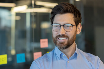 A man wearing glasses and a blue shirt is smiling in front of a wall with colorful sticky notes