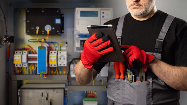 Man is electrician. Electrical equipment installer. Electrician with tablet computer in his hands. Cropped wizard sets up automation system. Man electrician in gray overalls and gloves