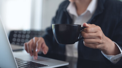 Close up of businesswoman hand holding a cup of coffee during working on laptop computer, surfing the internet at modern office, corporate business, online working