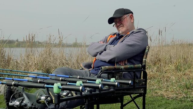 Mature adult senior angler fisherman sitting on chair sleeping, fishing rod setup with multiple rods on a stand with alarms set lines near lakeside in search of a catch, hobby leisure adventure
