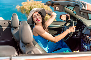 A woman is driving a red convertible with a hat on. She is smiling and she is enjoying the ride.