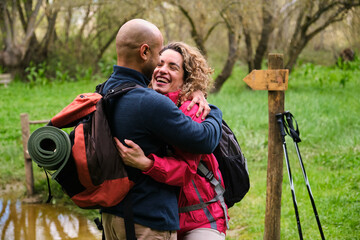A man and woman hug each other in a forest. They are laughing and wearing backpacks.