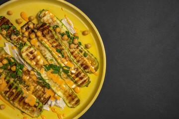 Grilled zucchini served with chopped parsley, chickpeas, tahini pasta, pink sauce on yellow platter over dark background.
