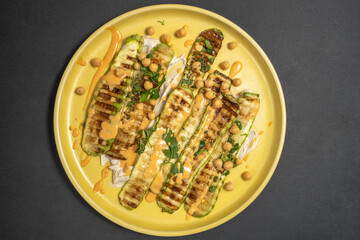 Grilled zucchini served with chopped parsley, chickpeas, tahini pasta, pink sauce on yellow platter over dark background.