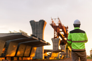 A construction worker is standing on a bridge, talking on his cell phone. The scene is set in a...