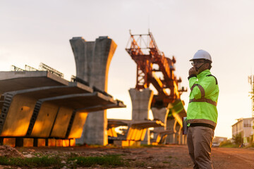 A construction worker is standing on a bridge, talking on his cell phone. The scene is set in a construction site, with a large crane in the background. The worker is wearing a yellow jacket