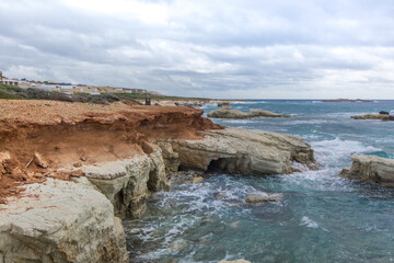 A rocky beach  with a cave positioned centrally. Waves crash against the shore, while the cave...
