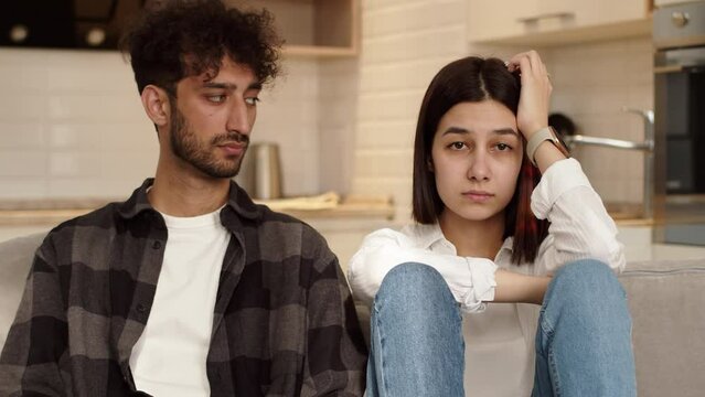 Young upset man sitting on a couch at home next to an offended young woman and looking at the camera after a quarrel. Concept of problems in couple relationship