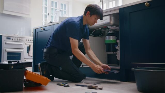 Male plumber with digital tablet fixing waste disposal unit in domestic kitchen  - shot in real time