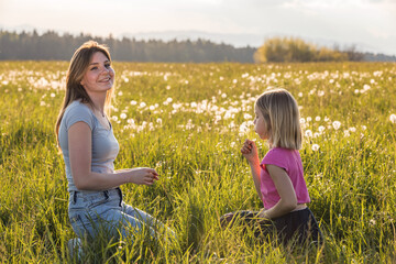 Mother and her little daughter blowing dandelions in the meadow on a sunny day. Moments of family happiness concept.