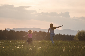 Joyful mother and her little daughter in the green meadow running with open arms at sunset. Life happiness concept.