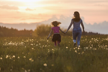 Mother and her little daughter running holding hands across the beautiful green grassland at sunset.