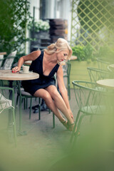 a sexy girl in a black dress at a table with coffee in a street cafe adjusts her shoe on her foot.
