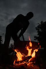 Silhouette of a man at dusk stirring food in a cauldron on an open fire.