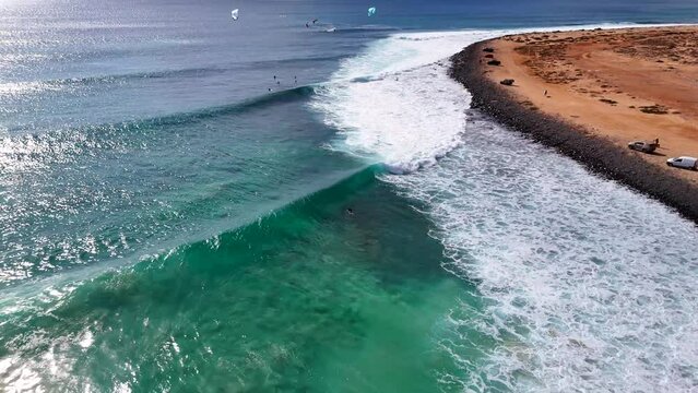 Flying over waves in Ponta Preta, Sal, Cape Verde, while a surfer paddles out. kitesurfers can be seen in the background. Camera pans up and reveals the coastline
