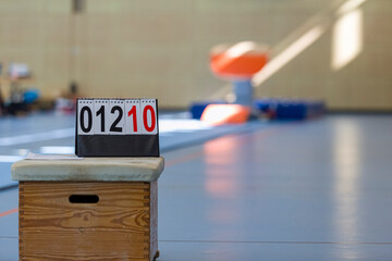 Scoreboard Wertungstafel vom Turnen auf einer Holzkiste in einer leeren Sporthalle, im Hintergrund ein Sprungtisch im Sonnenlicht, Zahlen zeigen 01210