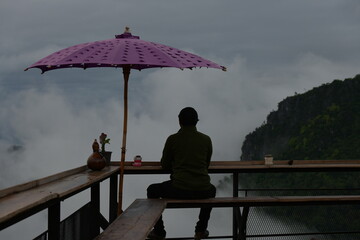Person under a purple umbrella sits on a platform high above clouds, contemplating the view....