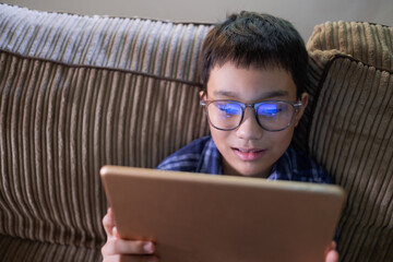 Young kid playing on his tablet close up, relaxing sitting on the sofa at home, browsing the digital online internet interweb, wearing glasses happy smiling, having an addiction problem.