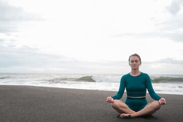 Fototapeta na wymiar Banner with woman in lotus position sits on a black sand beach and meditates against the backdrop of the ocean with big waves. Practice mindfulness and purity of mind. Concentration. Summer sports.