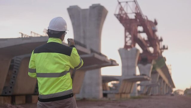 A construction worker is standing on a bridge, talking on his cell phone. The scene is set in a construction site, with a large crane in the background. The worker is wearing a yellow jacket