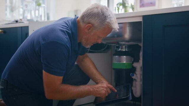 Mature male plumber with digital tablet fixing waste disposal unit in domestic kitchen  - shot in slow motion