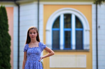 Fototapeta premium A young girl in a blue dress stands in front of a yellow building. The girl is smiling and looking at the camera. The scene is bright and cheerful, with the girl looking happy