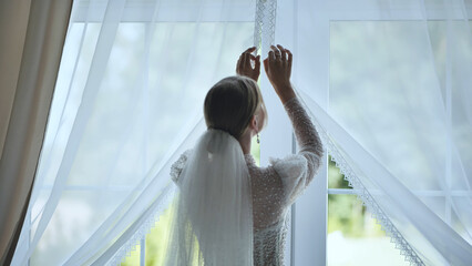 The bride adjusts the curtains at the window.