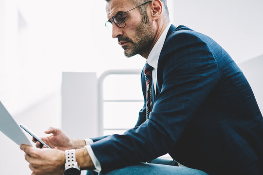 Concentrated mature entrepreneur dressed in formal wear reading accounting reports in text documents sitting in office.Pensive successful proud ceo with telephone in hands checking information data