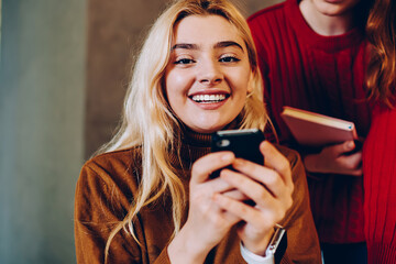 Cheerful hipster girls reading news about sales in web store on smartphone, happy teen girl showing...