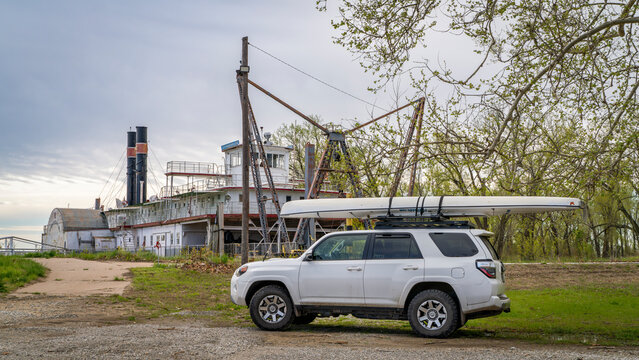 Brownville, NE, USA - April 21, 2024: Toyota 4Runner SUV with expedition canoe on a shore of Missouri River in spring scenery with a historic river dredge, Captain Meriwether Lewis, in background.