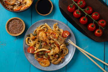 shrimp udon pad thai sauce gray plate on a blue table next to cherry tomatoes soy sauce and...