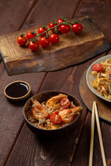 Rice with shrimp and udon in Pad Thai sauce on a wooden table next to cherry tomatoes, soy sauce and chopsticks