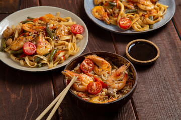 Rice with shrimp and udon in Pad Thai sauce on a wooden table next to cherry tomatoes, soy sauce and chopsticks