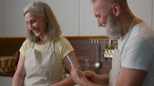 Caucasian married old senior couple happy family mature retired woman man cooking breakfast together in kitchen home cheerful smiling laughing husband hold mobile phone make photo wife cook food blog
