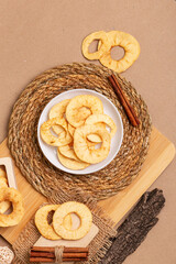 Crispy apple chips in a white round plate and in a wooden box, cinnamon sticks on a wooden desk from above on a beige background in rustic style. Healthy sweet snack.