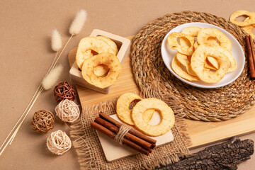 Crispy apple chips in a white round plate and in a wooden box, cinnamon sticks on a wooden desk on a beige background in rustic style. Healthy sweet snack.