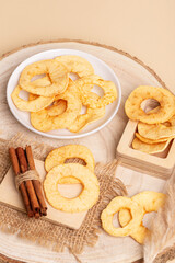 Tasty sweet apple chips in a plate and in a wooden box, cinnamon sticks on a wooden desk on a beige background.