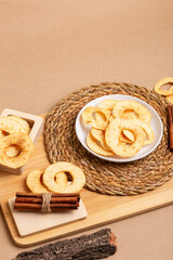Crispy apple chips in a white round plate and in a wooden box, cinnamon sticks on a wooden desk on a beige background in rustic style. Healthy sweet snack.