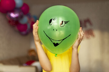 Green balloon with drawn smiley face in hands of girl in yellow dress covers her face of holiday at home